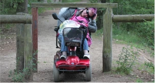 Une personne en fauteuil roulant électrique confrontée à une barrière sur un sentier, symbolisant les défis d'accessibilité et les obstacles rencontrés par les personnes en situation de handicap.